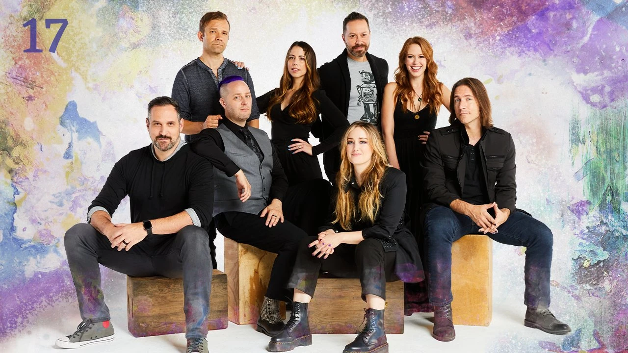 Thumbnail for episode 17. A group photo of Travis, Liam, Taliesin, Laura, Sam, Ashley, Marisha, and Matt sitting on large wooden blocks. They are dressed in black and gray and sit against a colorful backdrop with splashes of yellows, blues, purples, reds, and greens over white.