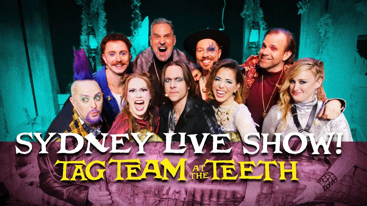 Thumbnail for “SYDNEY LIVE SHOW! TAG TEAM AT THE TEETH”. A group photo of Tom Cardy, Travis, Sam, Liam, Taliesin, Marisha, Matt, Laura, and Ashley posing on a stage next to each other looking towards the camera. The main cast are mostly dressed in the style of Chetney, Caleb, Ashton, Laudna, Jester, and Yasha. Tom wears a sparkly blue athletic jacket, Sam wears a Crocodile Dundee outfit, and Matt wears a black leather jacket and eye shadow. The stage is colorized in night vision green, and the bottom of the image is shaded maroon.