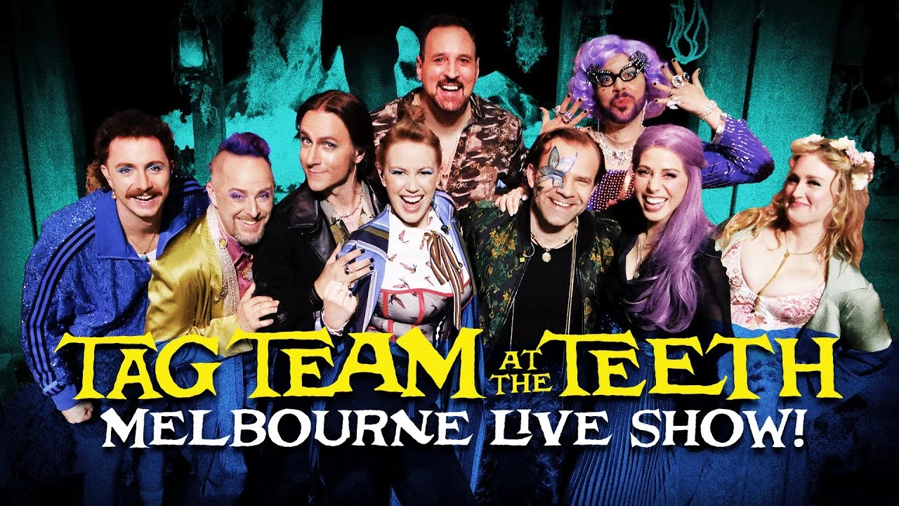 Thumbnail for “TAG TEAM AT THE TEETH. MELBOURNE LIVE SHOW!”. A group photo of Tom Cardy, Taliesin, Matt, Travis, Sam, Marisha, Liam, Laura, and Ashley posing on a stage next to each other smiling at the camera. The main cast are mostly dressed in the style of Caduceus, Beau, Fjord, Orym, Imogen, and Fearne. Tom wears a sparkly blue athletic jacket, Sam wears cat eye glasses, a purple wig, and rings, and Matt wears a black leather jacket and eye shadow. The stage is colorized in night vision green, and the bottom of the image is shaded blue.