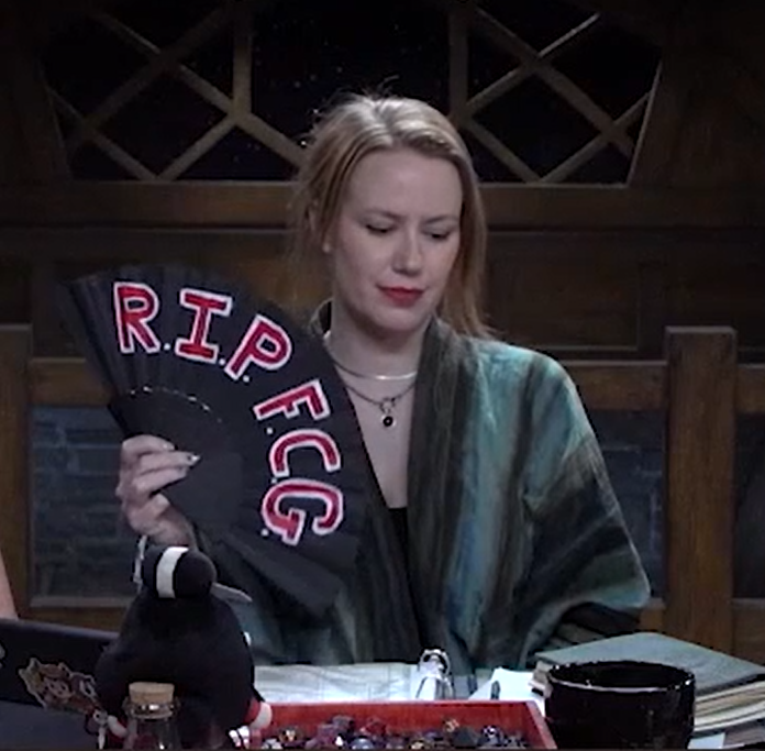 Marisha holding a black hand fan reading “R.I.P. F.C.G.” in red text outlined in white. She is looking down sadly.