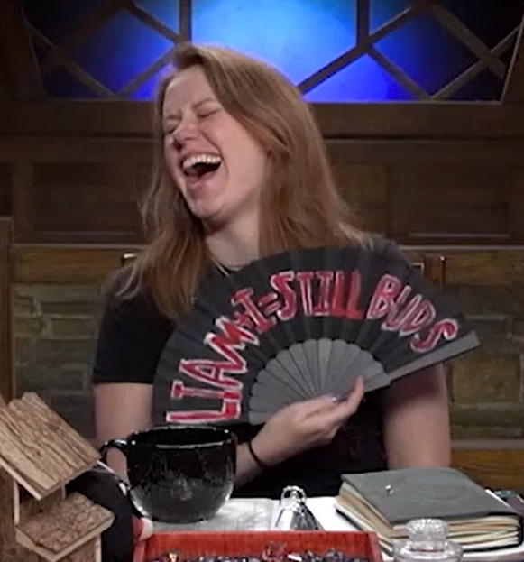 Marisha holding a black hand fan reading “LIAM + I = STILL BUDS” in red text outlined in white. Her head is craned to the side in uproarious laughter.