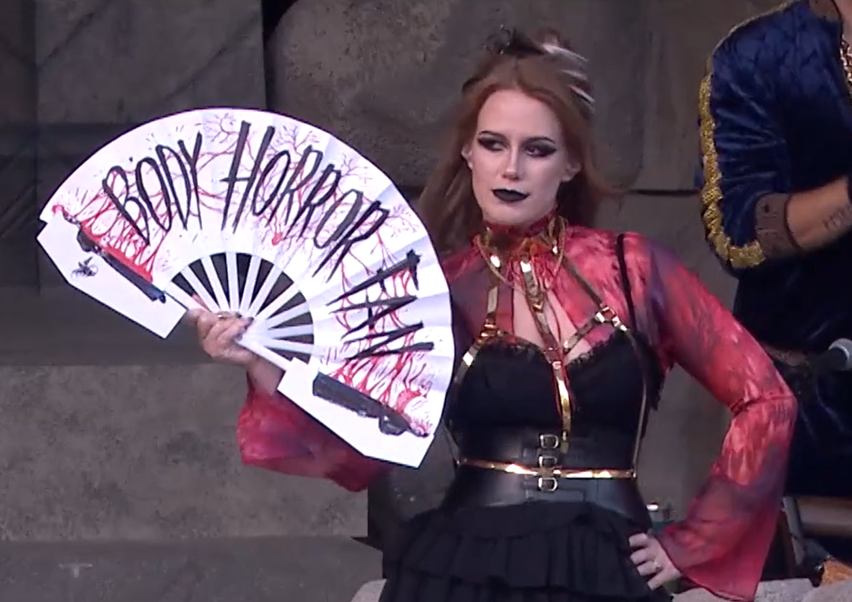 Marisha holding a white paper hand fan with the text “BODY HORROR FAN” in black writing connecting one side of Laudna’s body on the left of the fan with the other side on the other end. Blood connects Laudna’s two halves and spatters behind the lettering. Marisha is wearing a black dress with red sleeves and gold highlights and is standing posed with the fan open and her left hand on her hip.