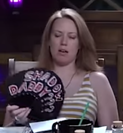 Marisha fanning herself with a black hand fan reading “SHADOW DADDY???” in red text outlined in white.