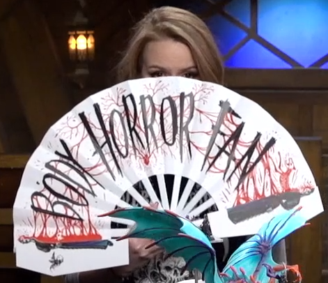 Marisha covering her face with a white paper hand fan with the text “BODY HORROR FAN” in black writing connecting one side of Laudna’s body on the left of the fan with the other side on the other end. Blood connects Laudna’s two halves and spatters behind the lettering.