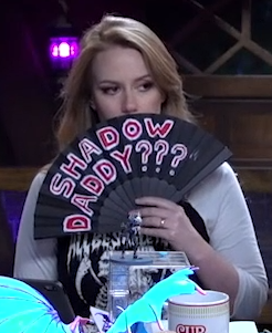 Marisha covering her face with a black hand fan reading “SHADOW DADDY???” in red text outlined in white.