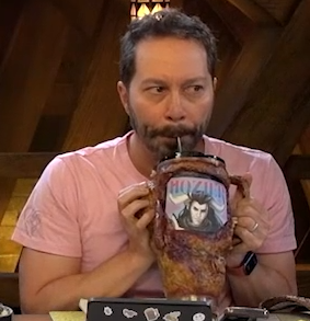 A tall cup covered in leathery monstrous skin. On the side is an image of Braius with the text “HOZIER” above it,  set on a pink and sky blue background. Sam is sipping from a straw placed in the top of the cup.