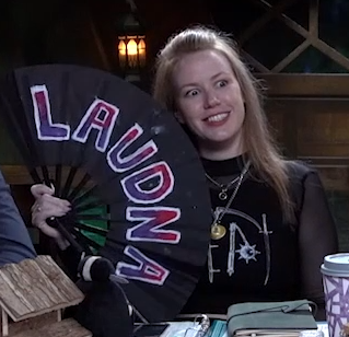 Marisha holding a black hand fan reading “LAUDNA” in purple and pink lettering outlined in white. She is making a creepy grin.