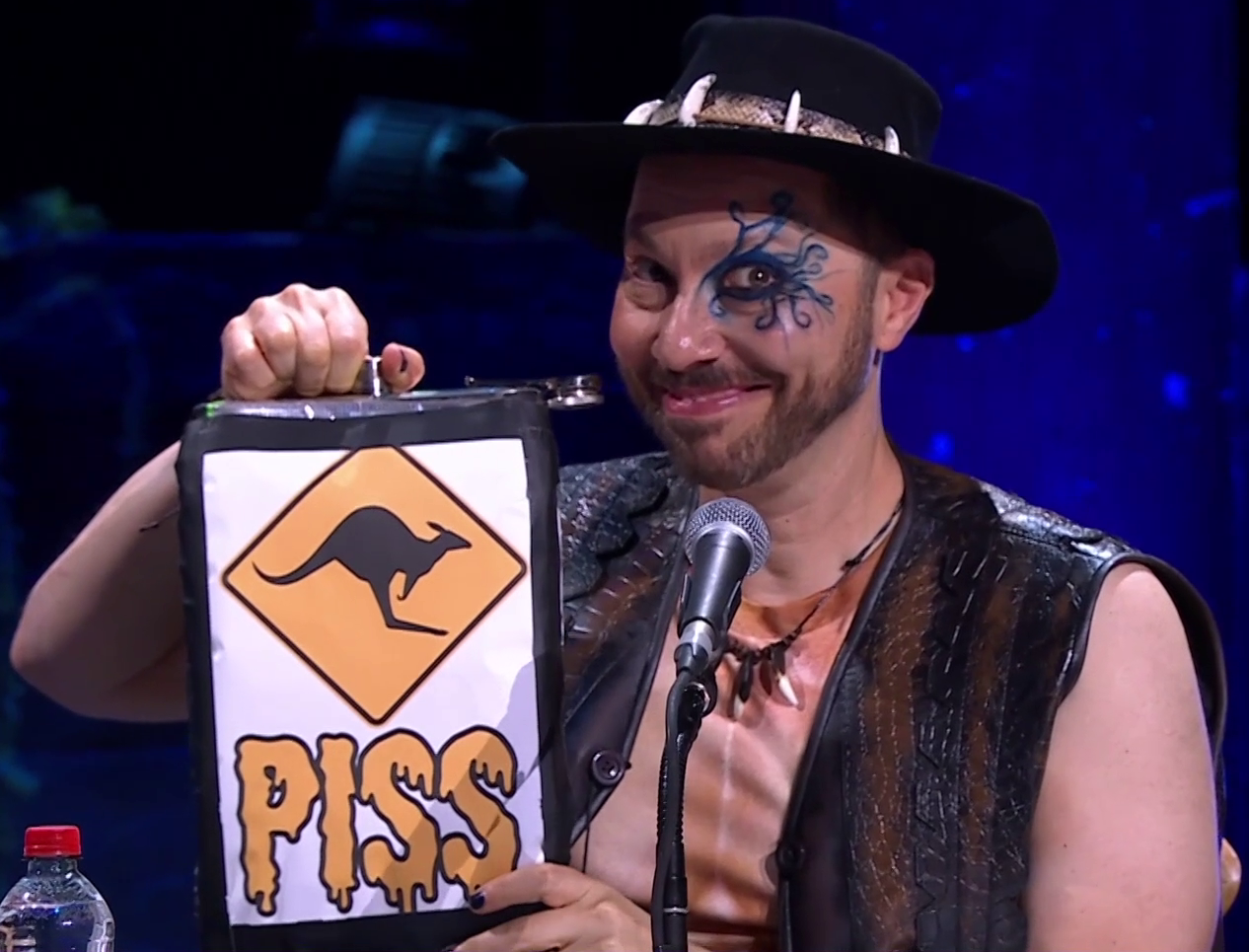 A comically-large flask. On the front is an image of a kangaroo-crossing road sign, underneath which is the text “PISS” in dripping yellow letters. Sam holds the flask, smiling at the camera. He wears a Crocodile Dundee outfit, complete with open crocodile-leather vest and black fedora ringed with crocodile teeth. Swirling blue paint is around his left eye.