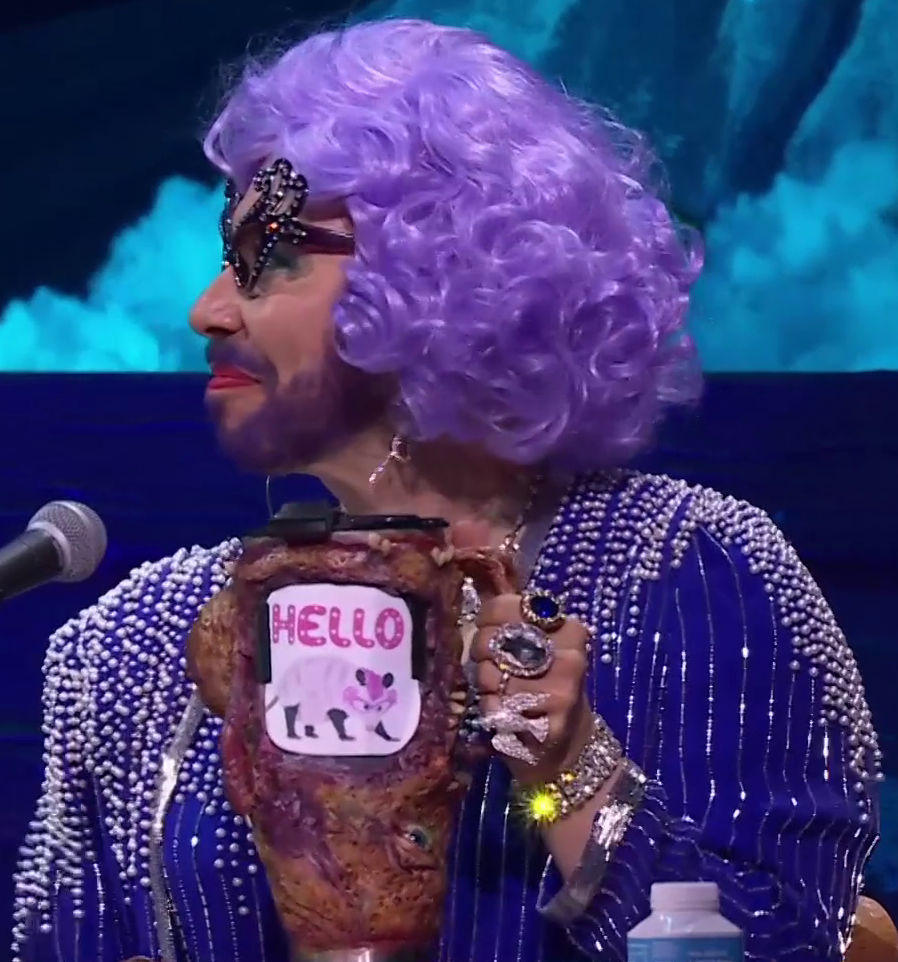 A tall cup covered in leathery monstrous skin. On the side is an image of a light pink possum under pink and white polkadot text reading “HELLO”. Sam holds the cup in his left hand. He wears a lavender curly wig, bedazzled cat eye glasses, lipstick, a blue pearled dress, and large jeweled rings on his hand. His beard is dyed purple. He looks to his right, smiling slightly.