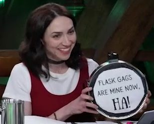 A circular canteen with a screw top. The text “FLASK GAGS ARE MINE NOW. HA!” is printed on a white sheet of paper taped to the front. Eden Riegel holds it, smiling.