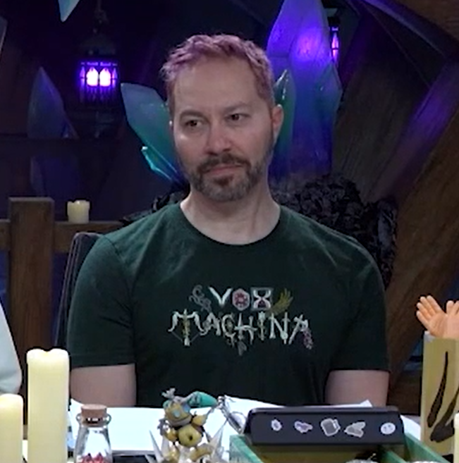 Sam wearing a forest green T-shirt that says “VOX MACHINA”. The letters are stylized, where for example the “O” is a d20, the “X” is an hourglass, the “M” is a bow and arrow, and the final “A” is an axe.