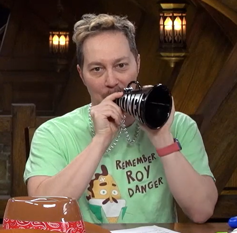 Sam wearing a seafoam green shirt with the text “REMEMBER ROY DANGER” next to a cartoon of a man with brown hair and mustache and cross bandage on his forehead. Sam is holding a small black megaphone that says “DONE!” in silver lettering.