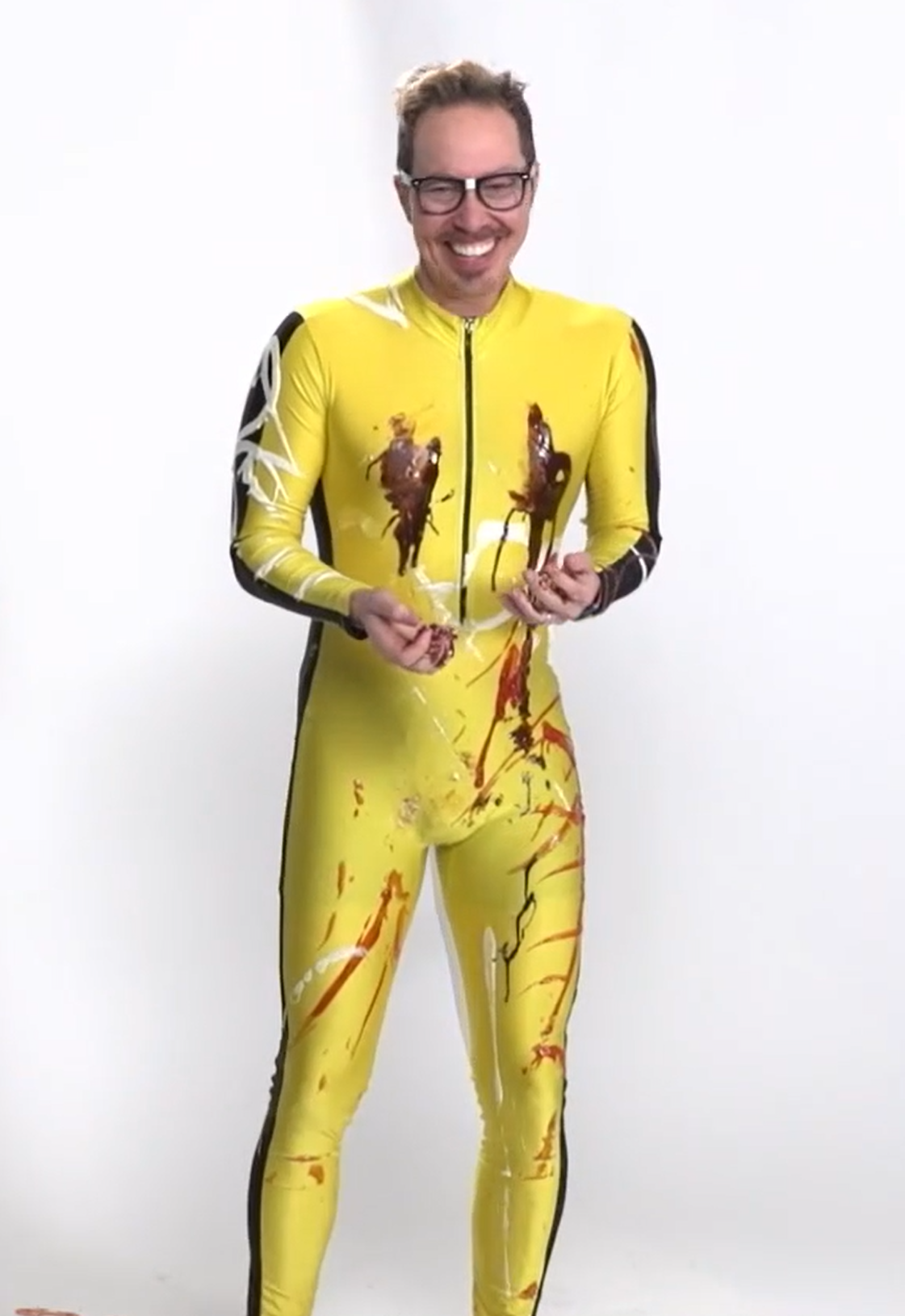 A full body image of Sam wearing a yellow jumpsuit with black stripes down the arms and sides. He is covered in streaks of various condiments such as ketchup, chocolate sauce, and mayonnaise. He is grimacing, his hands out in front of him.