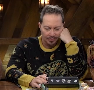 Sam wearing a black sweater with yellow around the collar, wrists, and elbows. The sweater is decorated with designs of stars, moons, bows and arrows, skyships, and tridents, with a tree bearing red fruit in the center of the chest.