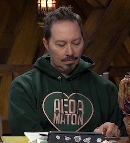 Sam wearing a green hoodie with a green heart bordered in rose gold on the front. Inside the heart is the text, also in rose gold, “AEORMATON”.