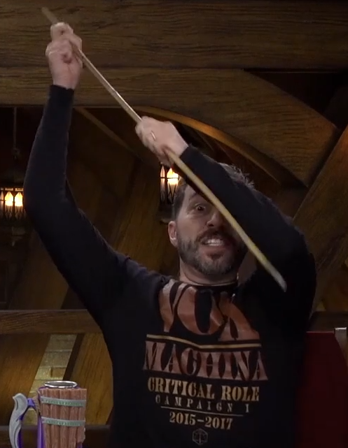 Sam wearing a black long-sleeve T-shirt. On it in dark orange and yellow bold font is the text “VOX MACHINA CRITICAL ROLE CAMPAIGN 1 2015–2017” with the Critical Role logo (a 2D representation of a d20) under the final line of text. Sam is pulling out a wooden blow dart gun and bearing his teeth menacingly.