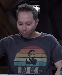 Sam wearing a gray T-shirt with a silhouette of Ruth Bader Ginsburg backed by colored stripes of red, orange, yellow, white, and green. The stripes form a shape similar to a rising or setting sun. Underneath the stripes are the letters “RBG” in large font. Sam’s cheeks are puffed out as if his mouth is full of water.