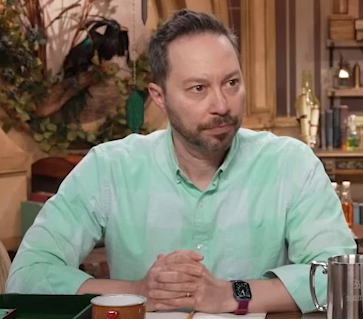 Sam wearing a pale green button-up shirt with the top button undone. Wide stripes of pale and slightly darker green form a checked pattern. He sits with his hands clasped on the table in front of him, leaning forward intently.