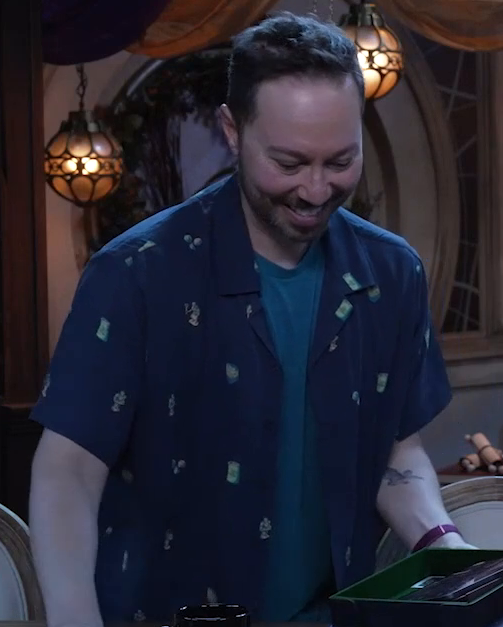 Sam standing at the table, wearing a navy blue button-up shirt unbuttoned to reveal a lighter blue T-shirt underneath. The button-up is sparsely decorated with small isdistinct images, including some cans.
