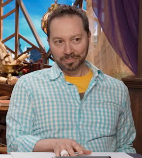Sam sitting at the table, wearing a light-blue and white checkered button-up shirt unbuttoned to reveal a yellow T-shirt underneath. The edge of an image is visible on the yellow shirt. Sam looks forward intently, his right hand placed palm-down on the table with a splint visible on his ring finger.