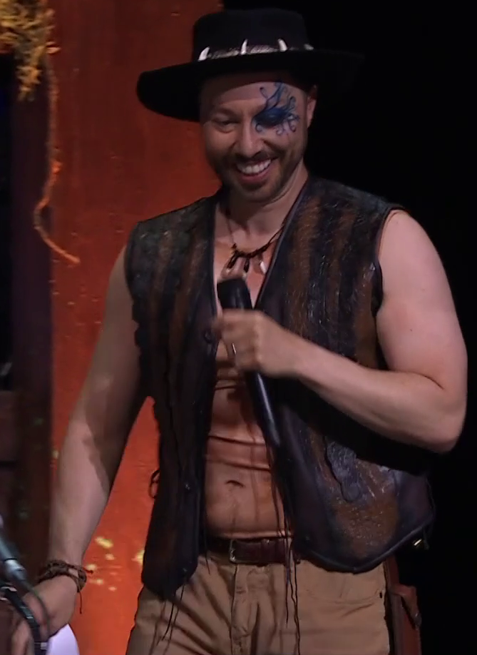 Sam wearing a Crocodile Dundee outfit. He has a crocodile-leather vest open to reveal a shirt displaying a fake muscular chest. A necklace with three crocodile teeth is around his neck and a black fedora ringed with crocodile teeth is on his head. He wears a messy leather bracelet on his right wrist, khaki pants, and a brown belt. Swirling blue paint is around his left eye. He smiles, holding a microphone and standing on a stage.
