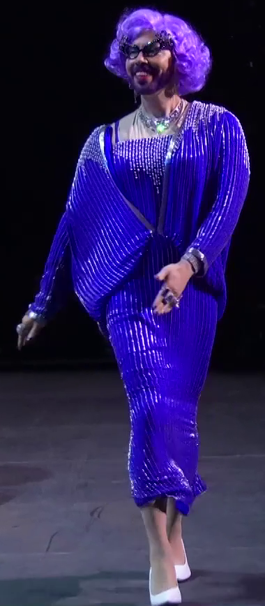 Sam strutting on a stage wearing a sparkly pearled blue dress and jacket. He has a curly purple wig, bedazzled cat eye glasses, and white high heels. A large ornate necklace is around his neck and his fingers are covered in large rings. Shiny silver bracelets adorn both his wrists. His beard is dyed purple.