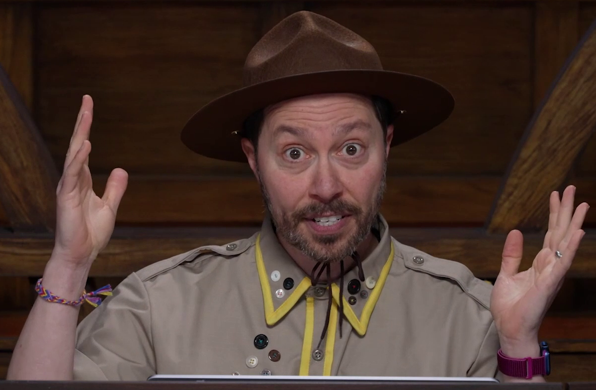 Sam wearing a tan scout uniform and brown brimmed campaign hat, with both hands raised and eyes wide mid speech. The uniform has yellow trim around the collar and along the central line of buttons. A number of other clothing buttons are attached haphazardly on the right breast and collar. The hat is strapped and tied around his chin. He wears a multi-color woven bracelet on his right wrist and a pink-banded watch on his left wrist.