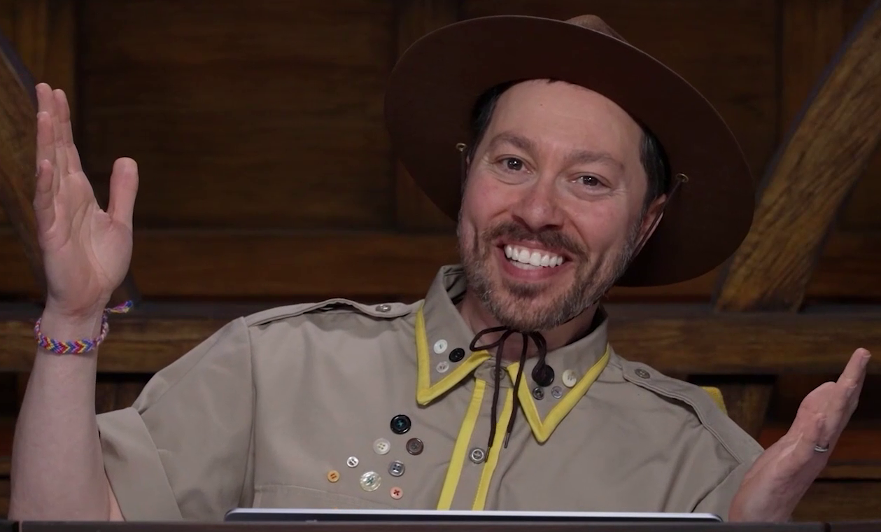 Sam wearing a tan scout uniform and brown brimmed campaign hat, with both hands raised and mouth open in a welcoming smile. The uniform has yellow trim around the collar and along the central line of buttons. A number of other clothing buttons are attached haphazardly on the right breast and collar. The hat is strapped and tied around his chin. He wears a multi-color woven bracelet on his right wrist.