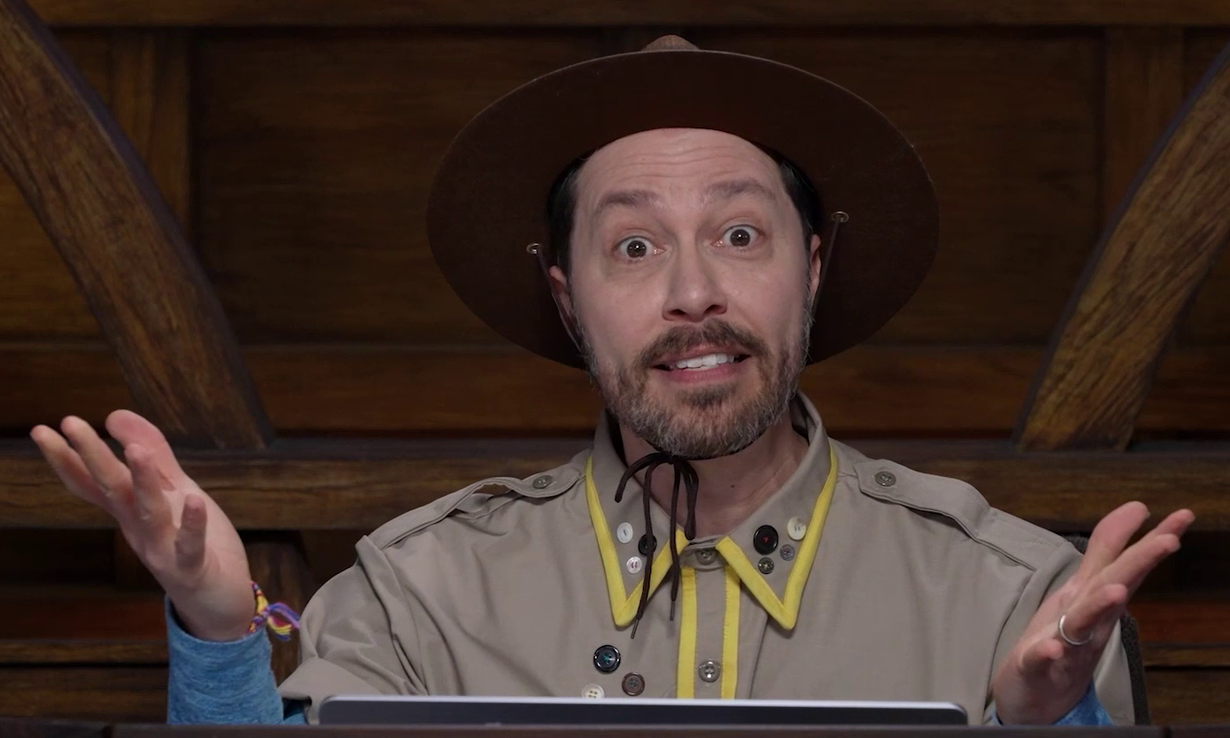 Sam wearing a tan scout uniform and brown brimmed campaign hat, with both hands raised in a goofy shrug. The uniform has yellow trim around the collar and along the central line of buttons. A number of other clothing buttons are attached haphazardly on the right breast and collar. The hat is strapped and tied around his chin. He wears a multi-color woven bracelet on his right wrist.