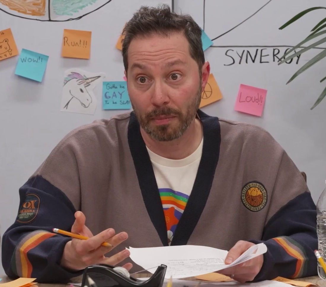 Sam wearing a roomy Vox Machina sweater over a white T-shirt bearing a rainbow, red star, and white flower. He holds a pencil and paper and seems to be engaged in some kind of serious discussion. The sweater is light gray with a dark blue collar and sleeves, and bands of red, yellow, gray, and green near the wrists. A patch on the right arm reads “VOX MACHINA. CRITICAL ROLL. CAMPAIGN 1. 2015–2017”. A patch on the left breast has the names of the Vox Machina members in a circle around the text “VOX MACHINA” in a bubble font. Behind him is a whiteboard with graphs, a drawing of a unicorn,  and a number of sticky notes bearing text like “Run!!!”, “Loud!!”, and “Gotta be GAY To be SLAY”.