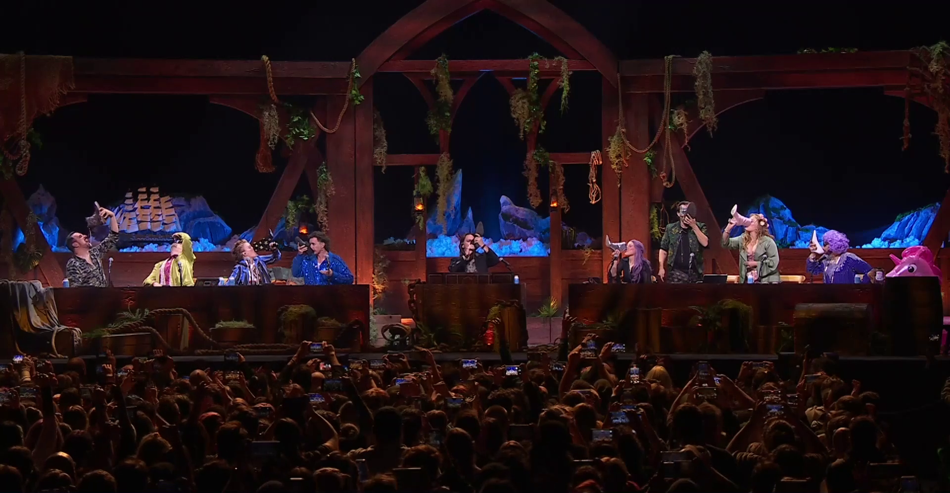 A wide shot of a stage where the cast of Critical Role are drinking from shoes. Travis lets liquid fall several inches from his boot onto his tongue, Taliesin tips back his head, Marisha drinks from a black boot with gold stars, Tom Cardy cautiously drinks from a black shoe, Matt tips back a black boot, Laura sips from a purple high-heeled boot, Liam stands and sips from a boot, Ashley stands and tips back a pink high-heeled boot, and Sam sips from a white stiletto. The audience all raises their phones to take pictures. The stage is set with a nautical design, including a model ship, lanterns, and rigging hung on a wooden backdrop. A pink inflatable unicorn is at the right side of the image next to Sam.