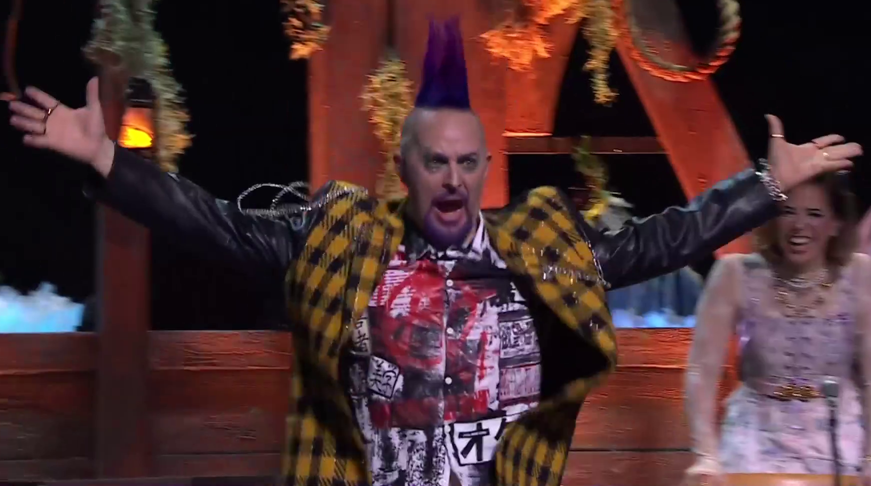 Taliesin dressed as Ashton raising his arms charismatically on a stage. He wears a yellow and black checkered leather jacket with black sleeves. Under the jacket is a black and white button-up shirt bearing Japanese characters and an anarchy symbol spray painted in red over the top of it. He has a tall purple mohawk and his beard is dyed purple. Chains bounce off the right shoulder and chest of his jacket. Laura dressed as Jester smiles in the background behind him.