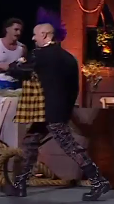 Taliesin dressed as Ashton striding across a stage. He wears a yellow and black checkered leather jacket with a black back and sleeves. He has a tall purple mohawk, black pants, and black combat boots. Chains drape off the top of the boots. Tom Cardy in a white tanktop is in the background.