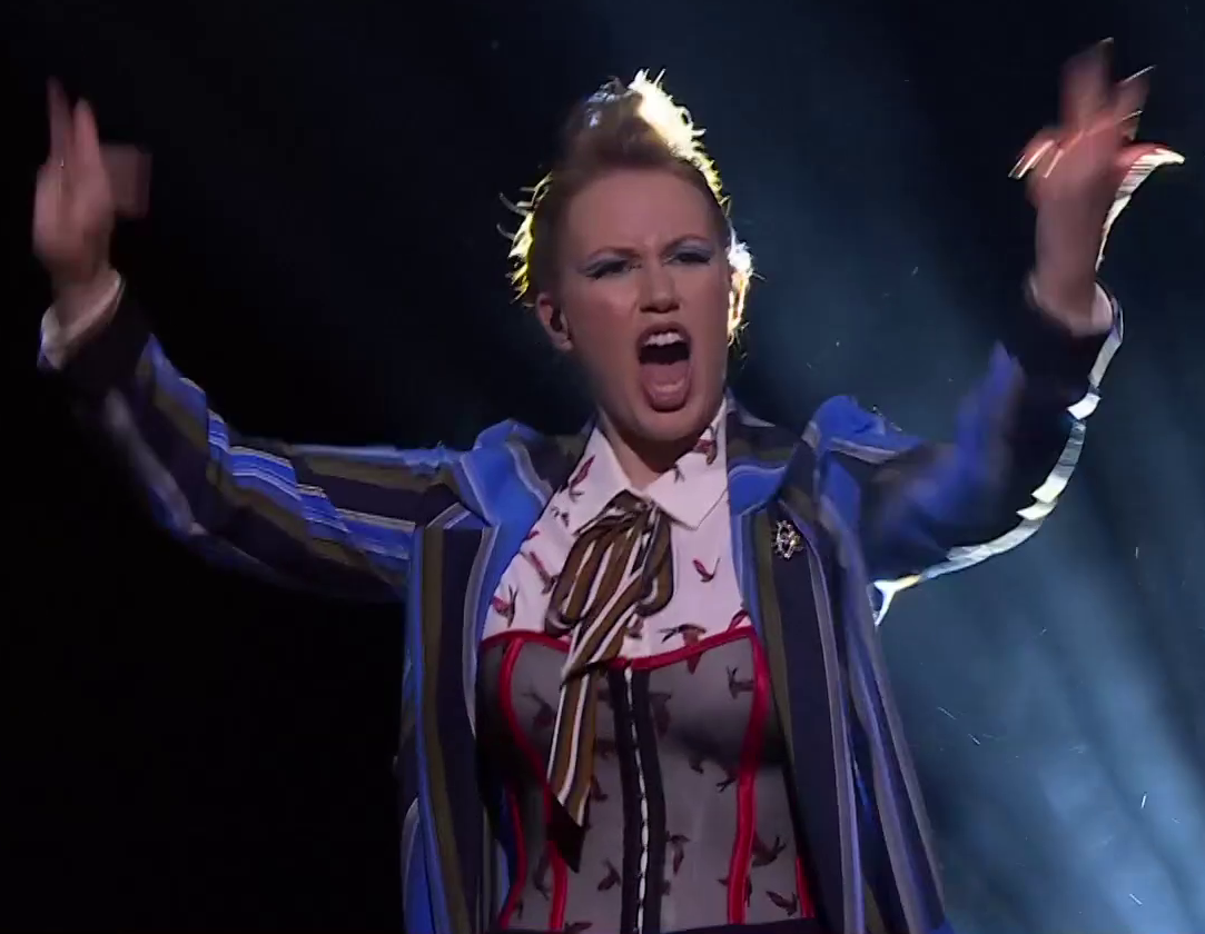 Marisha dressed as Beau on a stage, arms raised and charismatically pumping up a crowd. She wears a blue striped overcoat over a white collared shirt with birds on it. A mesh corset with red accents sits on top of the shirt. A brown striped bow is tied around her neck. An oval brooch reminiscent of an eye is on her jacket. An earpiece is in her right ear.