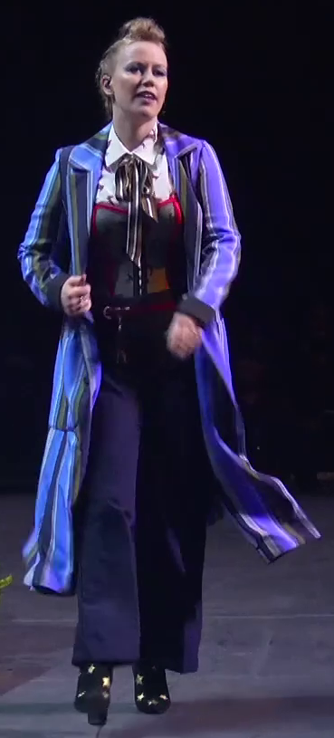 Marisha dressed as Beau striding across a stage. She wears a long blue striped overcoat billowing behind her over a white collared shirt with birds on it. A mesh corset with red accents sits on top of the shirt. A brown striped bow is tied around her neck. She has navy slacks and black high-heeled boots with golden stars on them. An earpiece is in her right ear.