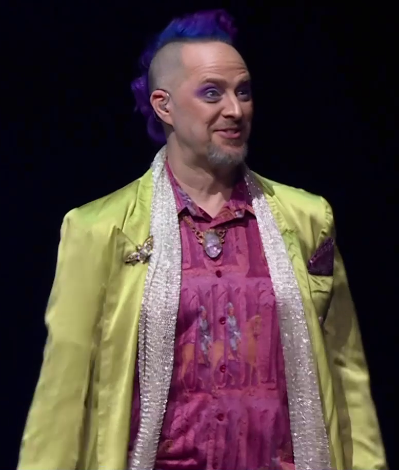Taliesin dressed as Caduceus standing on a stage, smiling. He wears a pink collared shirt under a yellow silk jacket and long white sparkly scarf. A large white crystal hangs around his neck, a brooch of a moth sits on his right lapel, and a purple hankerchief rests in his left jacket breast pocket. He has a purple mohawk and an earpiece in his right ear. The almost ethereal image of a person riding a horse is just visible on his shirt.