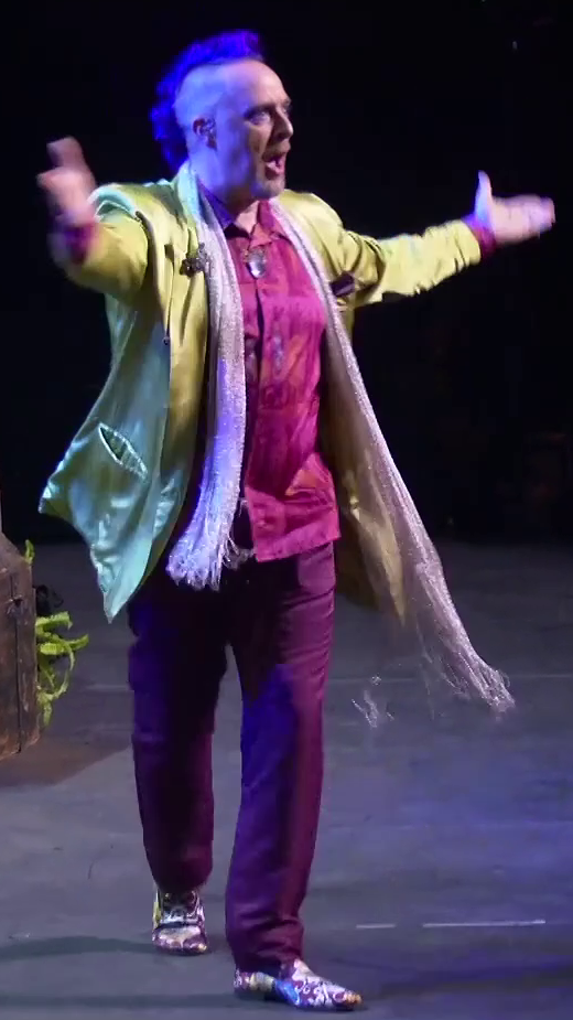 Taliesin dressed as Caduceus striding across a stage, hands raised to gesture for applause from an unseen audience. He wears a pink collared shirt under a yellow silk jacket and long white sparkly scarf. He has purple slacks and shoes bearing swirling colorful patterns. A large white crystal hangs around his neck, a brooch of a moth sits on his right lapel, and a purple hankerchief rests in his left jacket breast pocket. He has a purple mohawk and an earpiece in his right ear.