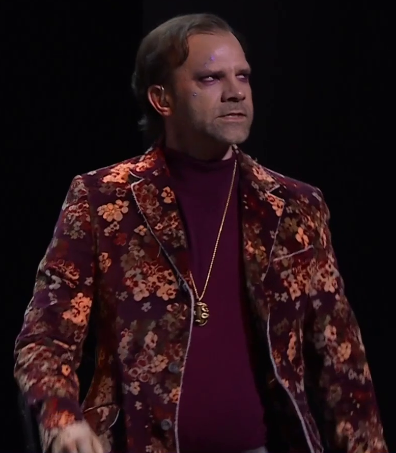 Liam dressed as Caleb on a stage, grimacing as he looks off into the distance. He wears a brown and maroon floral jacket open over a solid maroon shirt. A long gold necklace with a large pendant hangs down to his stomach. Purple eye shadow and some purple blemishes adorn his face. An earpiece is in his right ear.