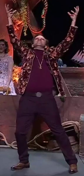 Liam dressed as Caleb on a stage, raising his arms as if summoning a spell. He wears a brown and maroon floral jacket open over a solid maroon shirt. A long gold necklace with a large pendant hangs down to his stomach. He has dark purple pants, a belt with a plain circular buckle, and shiny brown dress shoes. Laura dressed as Jester looks impressed in the background behind him.