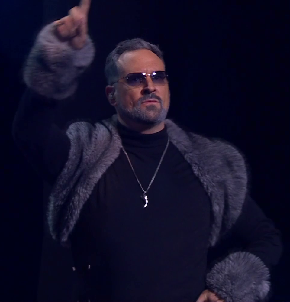 Travis dressed as Chetney standing straight on a stage, raising one finger as if pointing. He wears a gray fur shawl over a black turtleneck with gray fur cuffs. A silver necklace bearing a single curved tooth hangs over his chest. His eyes are visible behind dark shaded glasses. An earpiece is in his right ear.