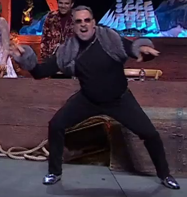 Travis dressed as Chetney posing in a ferocious crouch on a stage. He wears a gray fur shawl over a black turtleneck with gray fur cuffs. His black pants end in shiny silver shoes. His eyes are visible behind dark shaded glasses. Liam dressed as Caleb smiles in the background behind him, and Laura dressed as Jester is next to him, partially out of frame.