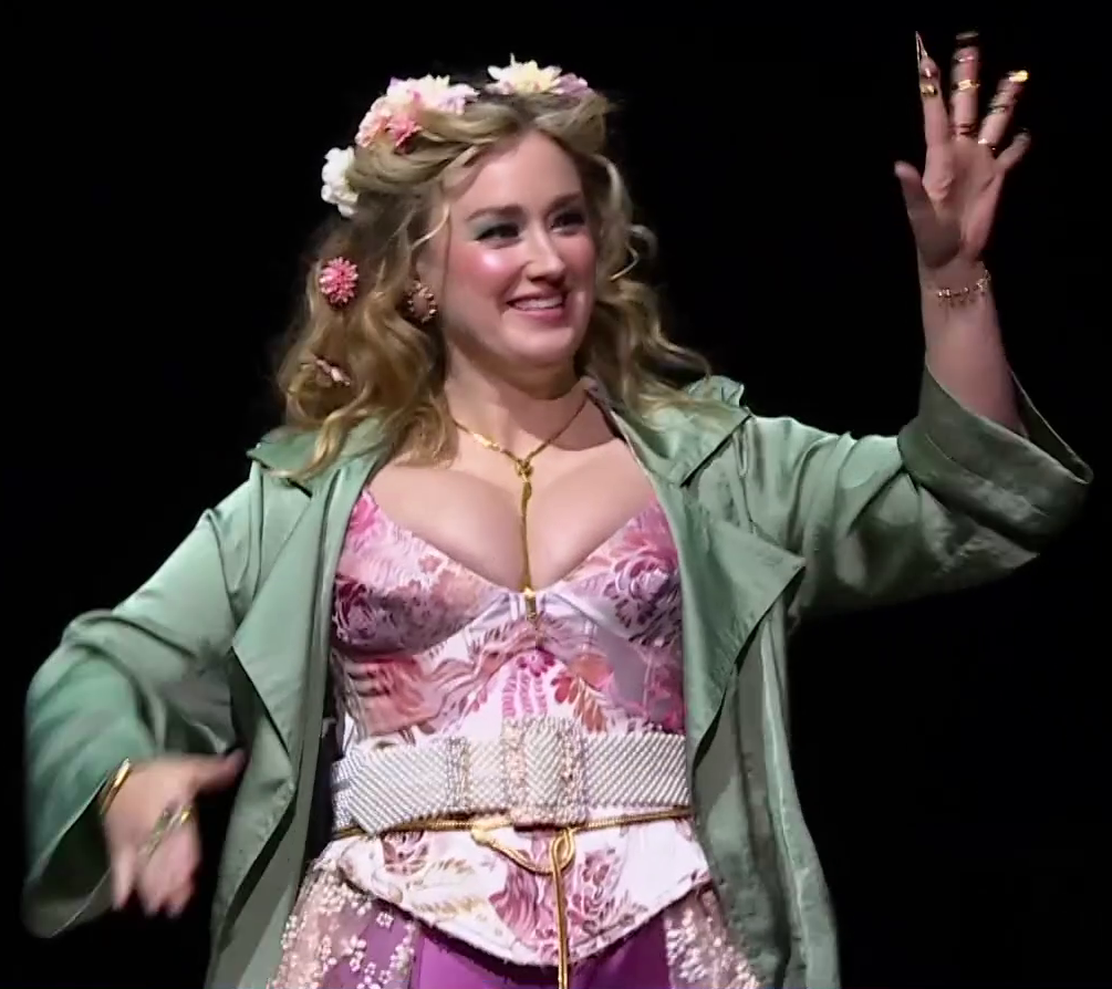 Ashley dressed as Fearne on a stage, smiling and waving at an off-screen crowd. She wears a floral pink, low-cut corset top under a seafoam green overcoat. Yellow, white, and pink flowers are strewn in her wavy brown hair. A white beaded belt sits around her waist. A golden cord hangs around her neck, and another one is tied just below the belt. Golden spiky tips are attached to her left hand’s fingers.