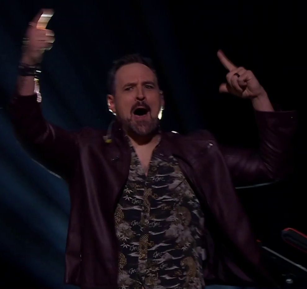 Travis dressed as Fjord on a stage, pointing and yelling to pump up an off-screen crowd. He wears a dark-tone collared shirt with images of tigers under a maroon leather jacket.