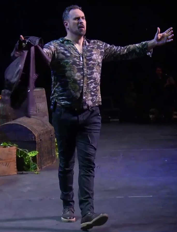 Travis dressed as Fjord striding across a stage, arms open towards an off-screen audience. He wears a dark-tone collared shirt with images of tigers and holds a maroon leather jacket in his right hand. He has black slacks, brown work boots with white soles, and a sound pack on his waist.