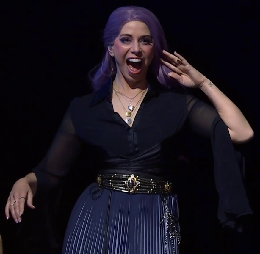 Laura dressed as Imogen on a stage, smiling and brushing back her long purple hair. She wears a black top with sheer sleeves tucked into a bronze metalic belt with an ovular gem set in the front and chains hanging off it. She has three necklaces, one a silver locket in a shape like a heart, the other two gold and solver circles pierced through their centers. A pleated purple skirt emerges from the belt. There are rings on her right hand.