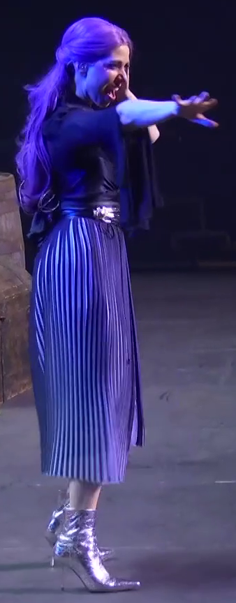 Laura dressed as Imogen on a stage, left hand to her temple and the other outstretched as if casting a spell. She wears a black top tucked into a metalic belt. A pleated purple skirt emerges from the belt, reaching to her mid calves. She has shiny silver high-heeled boots.