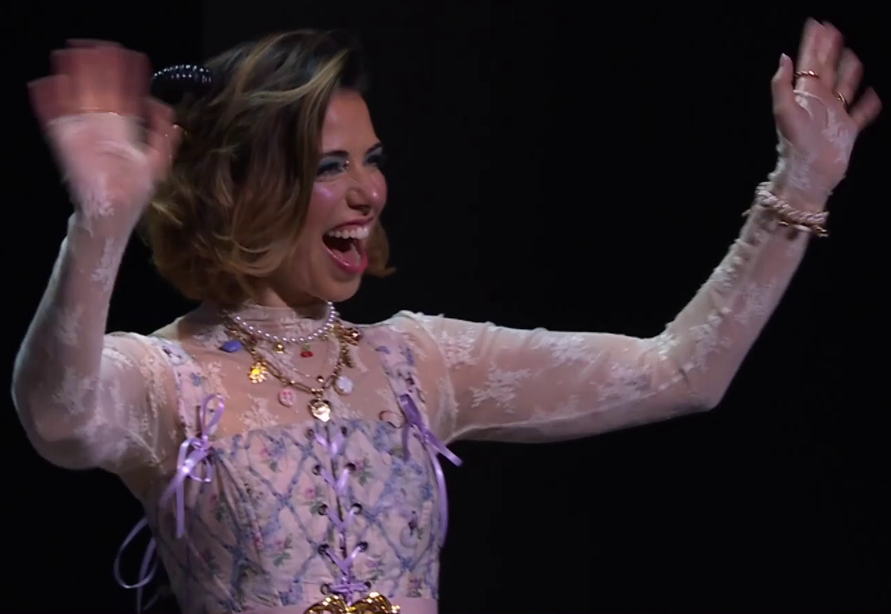 Laura dressed as Jester on a stage, smiling and waving at an off-screen crowd. She wears a lacy white and purple floral top whose bodice is tied with purple ribbons. She has two necklaces, one pearls and the other gold with shiny charms such as buttons. Black horns curl out from her brown hair. Rings adorn her fingers and a white pearly bracelet is on her left wrist. A gold belt bucket is just visible at the bottom of the image.