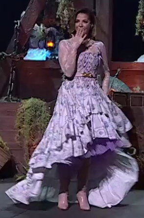 Laura dressed as jester on a stage, her hand placed over her mouth in mock surprise. She wears a lacy white and purple layered dress which billows out at the bottom. She has pink high heels and a gold belt.