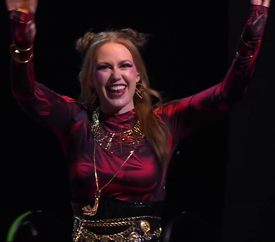 Marisha dressed as Laudna on a stage, smiling and waving at an off-screen crowd. She wears a red silk top with a geometric gold necklace and triangular earrings. A longer necklace bearing golden tassels at the end hangs above her stomach. A black corset-like belt with elaborate gold chains and adornments wraps around her waist. Her hair is mostly down, with two buns at the top of her head.
