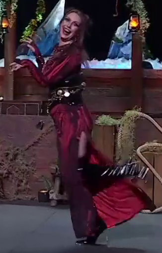 Marisha dressed as Laudna on a stage, posing with one leg up making a creepy face. She wears a red silk top slitted dress under a black corset-like belt with elaborate gold chains and adornments. She has tall black high-heeled boots.